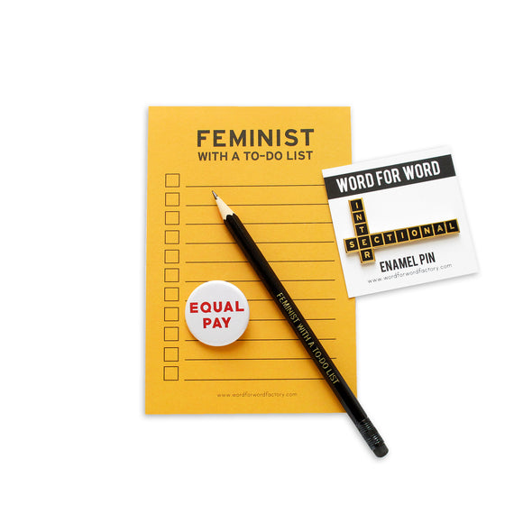 Feminist Collection of Products including a yellow Feminist With A To-Do List Notepad, And Equal Pay Pinback Button, an Intersectional Enamel pin, and a black Feminist With A To-Do List Pencil