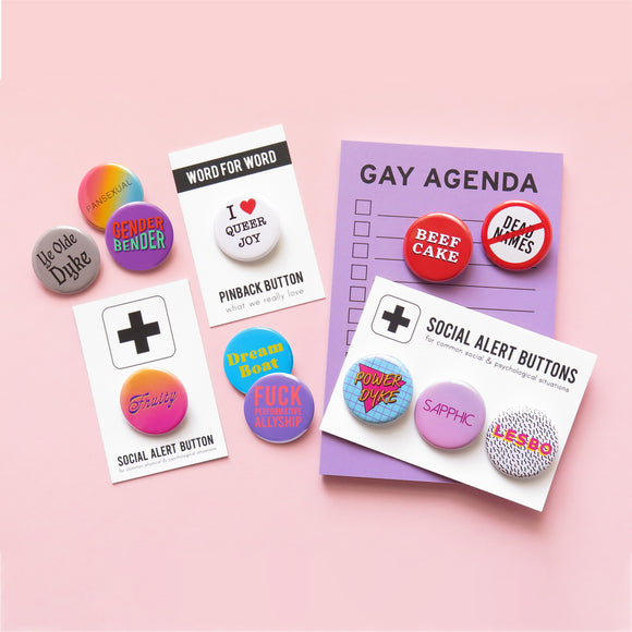 Collection of queer goods, it includes a purple Gay Agenda notepad, a purple Gay Agenda pencil, a Queer enamel pin, a pink Queer Femme pinback button, and a green multi-colored Gay Was pinback button