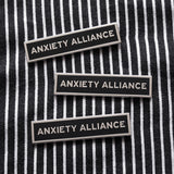 Three thin rectangle enamel pins that read ANXIETY ALLIANCE in silver text with black enamel. Pinned to a black & white vertical stripped jacket.