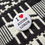 Round white pinback button that reads I Love Audre Lorde, with love indicated with a red heart. Pinned to a black and white woven jacket.