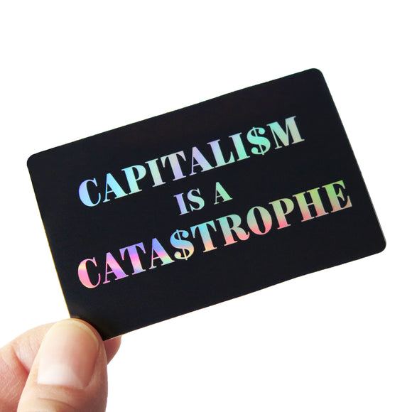 A black rectangular sticker with rounded corners that reads CAPITALISM IS A CATASTROPHE in holographic text, over 3 lines. The S's are replaced with dollar signs. Held in the lower left hand corner by a thumb & forefinger.