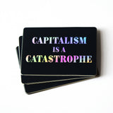 a stack of black rectangular stickers with rounded corners that reads CAPITALISM IS A CATASTROPHE in holographic text, over 3 lines. The S's are replaced with dollar signs. 