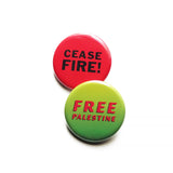 Two overlapping pinback buttons. The round button in the forefront is a neon green ombre that reads FREE PALESTINE in red text. The one behind is a round red pinback button that reads CEASE FIRE ! in black text. 