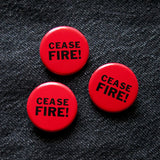 Three round red pinback button that reads CEASE FIRE! in two lines in black text. The buttons are pinned to black denim. 