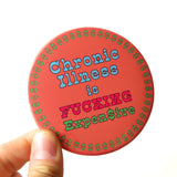 Large coral colored pinback button with an ring of green dollar signs around the edge of the button, reads CHRONIC ILLNESS IS in baby blue, FUCKING in hot pink, EXPEN$IVE in green