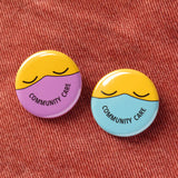 Two round buttons with a yellow emoji faces with closed eyes, on the left face is a lavender mask that reads COMMUNITY CARE in the shape of a smile. One the right face is a mint green mask that reads COMMUNITY CARE in the shape of a smile. Buttons are on a red-orange denim background.