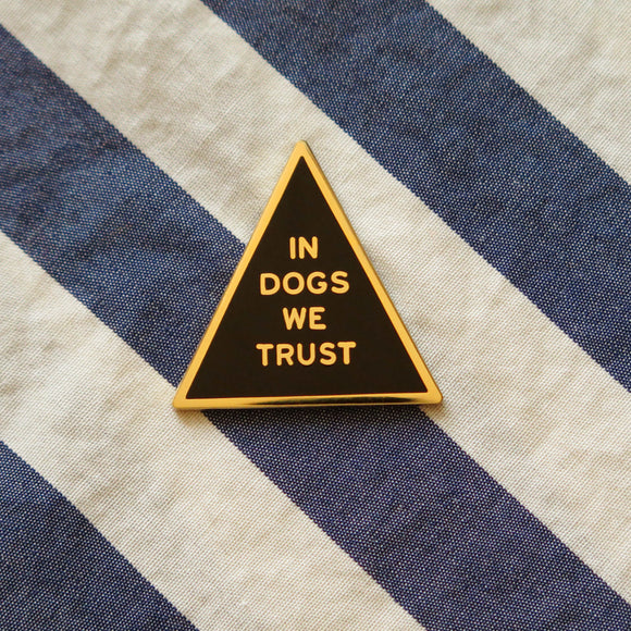 Triangle shaped gold & black enamel pin that reads IN DOGS WE TRUST. Gold text and outline on a black enamel background.