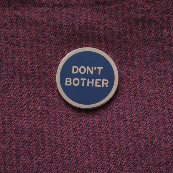 Round navy blue enamel pin that reads DON'T BOTHER in silver text and outline. Pinned to maroon striped denim.