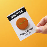 A orange-brown pinback button that reads TOAD in light green text. Button is on a black and white backing card that reads PERFECT PAIR, Pinback Button Share one
