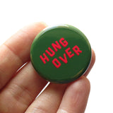 A round dark green pinback button that reads HUNG OVER in slightly askew neon red letters for a Christmas look.  Held in a hand, up close.