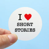 A round white sticker held in the corner with a thumb & forefinger against a light blue background. Sticker reads: I LOVE SHORT STORIES with love being represented by a red heart.