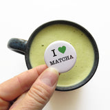 A round white pinback button that reads I LOVE MATCHA, has a green heart representing love, and the rest of the phrase is in a classic serif font.  Button is held over a mug of green matcha latte.