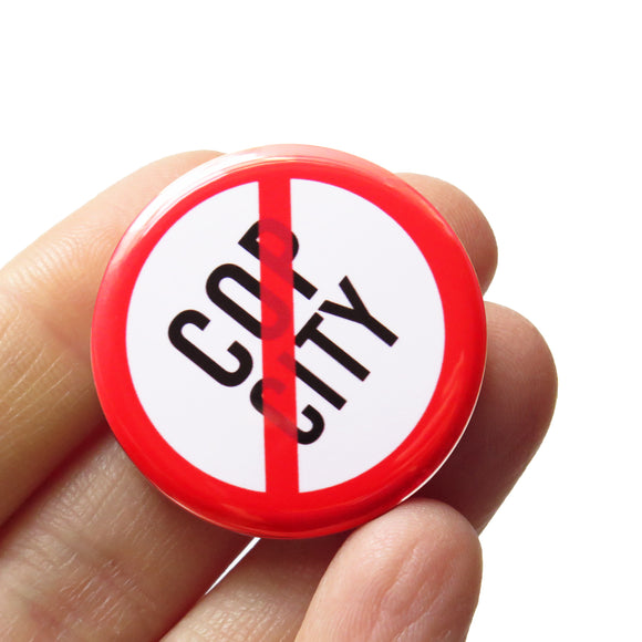 A round white button that reads stop COPY CITY in black text with a red circle and slash to indicate being against cop city. Button badge is held in a hand.
