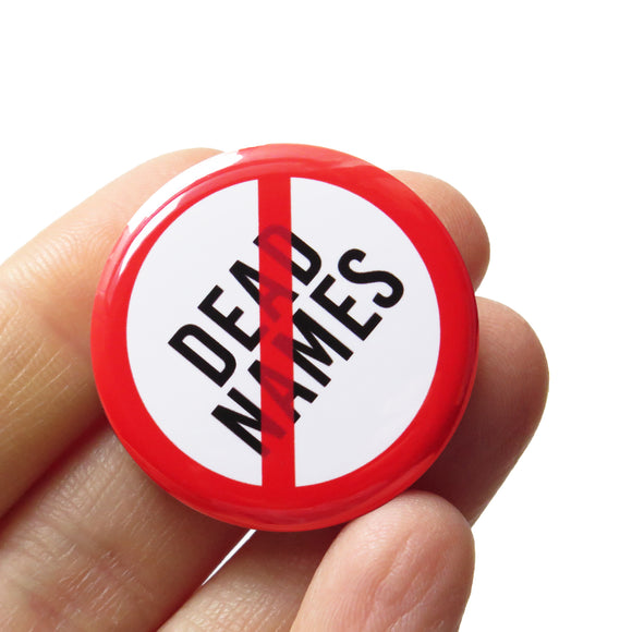 A round white button that reads NO DEAD NAMES in black text with a red circle and slash to indicate being against using dead names when referring to trans folks. Badge is held in a hand.