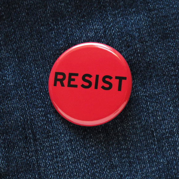 A round bright red pinback button that reads RESIST in black type. Button is on blue denim