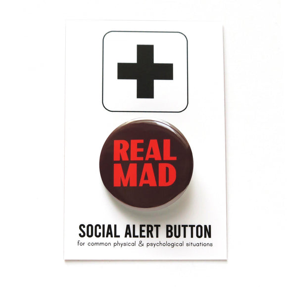 A dark brown pinback button with thick lava orange text that reads REAL MAD in two lines taking up all the space on the face of the button. Button badge is on a Social Alert Button backing card.