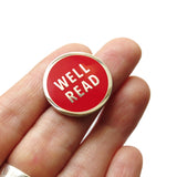 Red enamel lapel pin that reads WELL READ in silver metal. Pin is held in a hand, but comes on a branded Word For Word Factory backing card.
