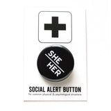 Round pinback button with black background which reads SHE/HER on an angle in white text, in a san serif font. On a white backing card, black plus sign at the top. Below reads: Social Alert Button, with tiny text beneath it which says, for common physical & psychological situations