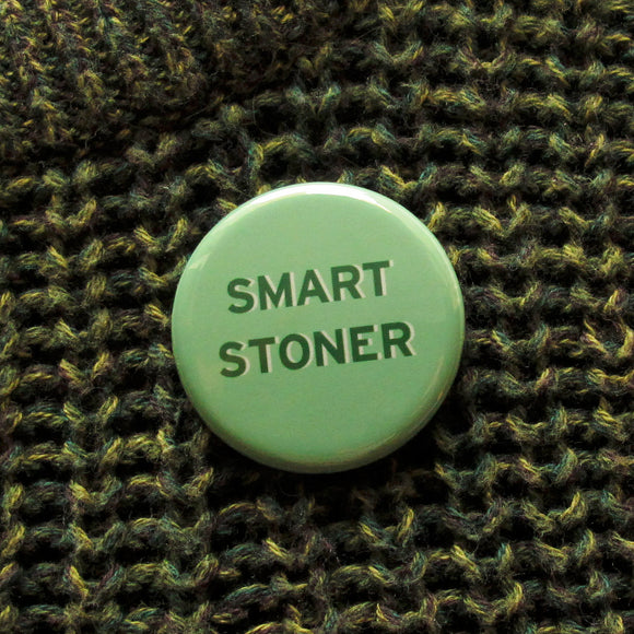 Round pinback button that says SMART STONER. Dark green text on a light green background. Button is pinned to a olive green knit sweater.