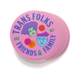 A stack of round pink sticker that reads TRANS FOLKS ARE MY FRIENDS & FAMILY around the permitter of the sticker in periwinkle & green text, with two illustrated faces in the middle, one with minty skin, purple hair and pink lips. The other with blue skin, glasses and a wavy swoosh of magenta hair. Little pink, orange & yellow flowers are decorating the negative space around them. 