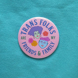 Large light-pink round pinback button, reads TRANS FOLKS ARE MY FRIENDS & FAMILY in blue & green text around the edge of button. Center has illustrated drawings of 2 heads with little flowers in the negative space. Head on the left is feminine with mint green skin, voluminous lavender hair, hoop earrings & pink lipstick. The face on the right is masculine with square jaw, light blue skin, round purple glasses, wavy pink hair, chin dimple & small smile. The button is on a a turquoise sweatshirt