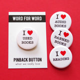 Four round white I heart pinback buttons that read: I love Used Books, I Love Audio Books, I Love Banned books,  I Love Reading. All on a red background, one book on a Word For Word branded backing card.
