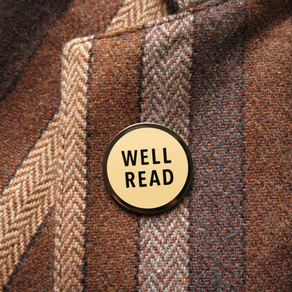 Round cream enamel pin that reads WELL READ in gunmetal dark silver text. Pin is on the lapel of a brown tweed blazer.