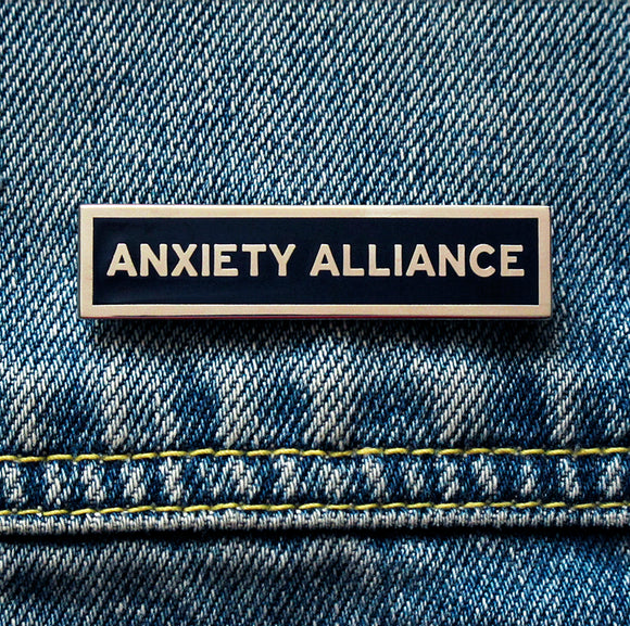 A thin rectangle enamel pin that reads ANXIETY ALLIANCE in silver with a black background and silver metal outline. Pinned to a blue denim jacket.