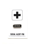 A capsule shaped enamel pin that reads DYKE in silver sans serif font on a black background. Pin is on Social Alert Pin backing card with a plus sign at the top.