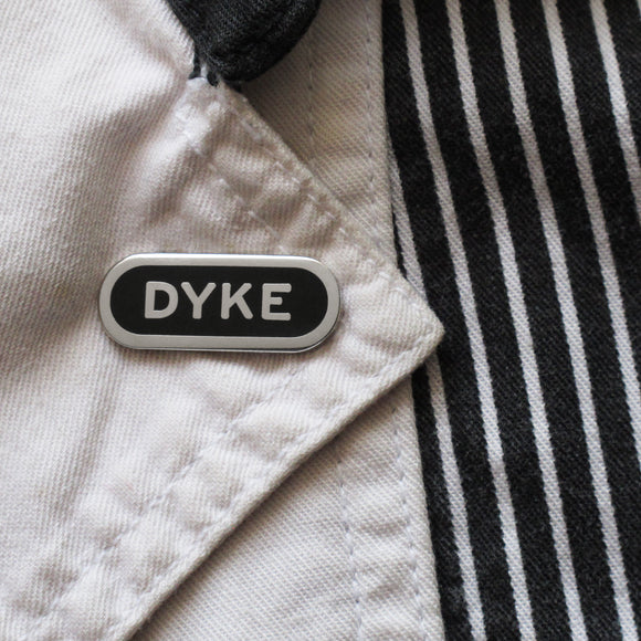 A cute queer capsule shaped silver and black enamel pin that reads DYKE, pinned to a black & white jacket, with a white lapel. Fun lesbian gift.