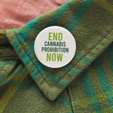 A round white pinback button that reads End Cannabis Prohibition Now, in light & dark green text.  Badge is pinned to a green plaid flannel shirt lapel.