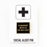 Gold & black rectangle enamel pin that reads FEMINIST WITH A TO-DO LIST.  Pinned on a Social Alert Pin branded backing card.