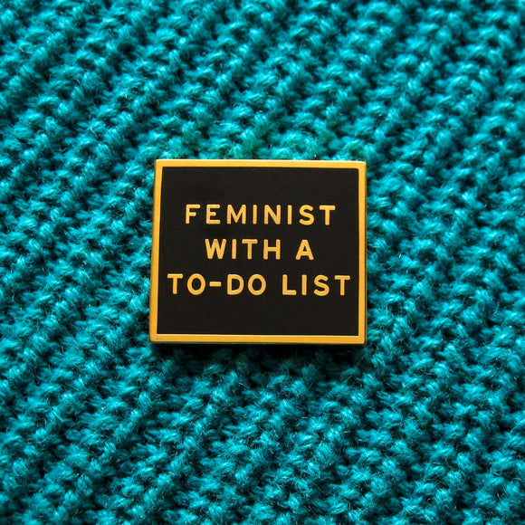 Gold & black rectangle enamel pin that reads FEMINIST WITH A TO-DO LIST. Pinned to a teal blue knitted sweater.