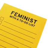 Close-up of the top part of a lined notepad with checkboxes that says  FEMINIST WITH A TO-DO LIST at the top in black text.   The paper is goldenrod yellow color.