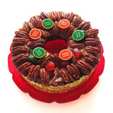 A classic nutty fruitcake on a vintage red plate, with glazed pecans & gold and red and gold and green round enamel pins scattered along the top like fruit cake jellies.
