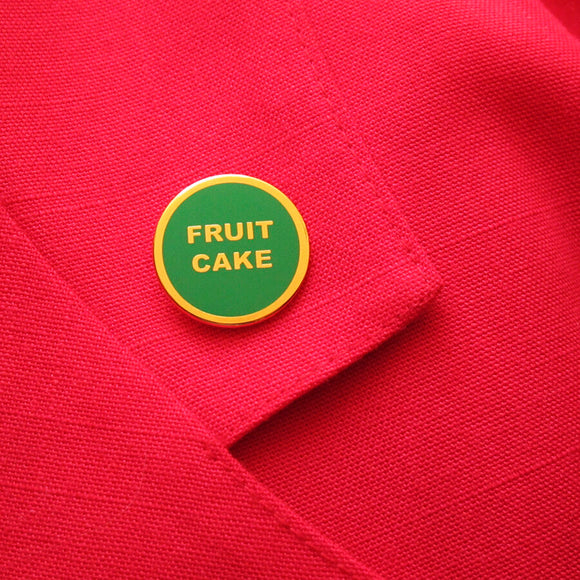 small circular enamel pin, green enamel background with gold text that reads FRUIT CAKE across two lines. Gold colored outline. Pin is on a red blazer lapel.