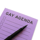 Close Up of a lavender notepad that reads GAY AGENDA in bold black text at the top, with checkboxes on the left side, and lines to the right, to write your to-do list! A black felt tip pen lays across the notepad. 