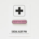 Capsule shaped enamel pin that says HEARTBREAKER.  Silver text and outline on a pink enamel background on a Social Alert Pin backing card.