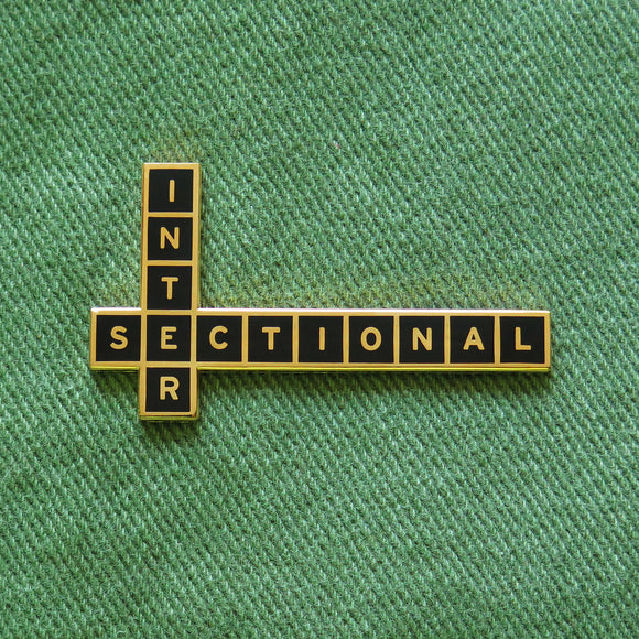 A gold & black enamel pin that looks like a tiny cross word two intersecting words that reads INTER SECTIONAL.