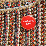 Shiny bright red pinback button with white san serif test reading, Migraine Day.  The button is pinned to a multicolor knit sweater with flecks of cream, black, brown read and pale blue.