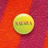 Pinback button with yellow to green ombre background, with pink to green ombre text that reads NAUSEA in a wavy serif font. Button is on a bright orange and magenta Terry cloth robe.