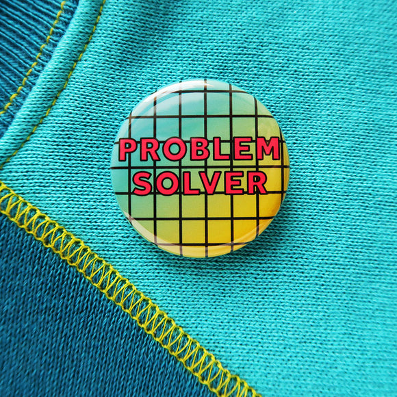 A round pin back button that reads PROBLEM SOLVER in dark pink text over a grid pattern with a blue to yellow gradient in the background. Button is on a blue aqua sweatshirt with yellow stitching.
