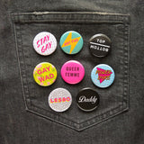 Black jeans back pocket with a bunch of bright, round gay & queer pinback buttons pinned on. Featuring Stay Gay, Art Fag, Top/Bottom, Gay Wad, Queer Femme, Power Dyke, Less, Daddy
