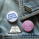 A old denim jacket with a mix of political pinback buttons & enamel pins reading: Queer, The Personal Is Political, Equal Rights Matter & Trans Rights Are Human Rights.