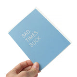 Hand holding a powder blue sympathy card that says SAD TIMES SUCK