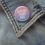 Light pink & blue, round, pinback button that reads SUPPORT TRANS KIDS. Button is pinned to the collar of an old denim jean jacket.