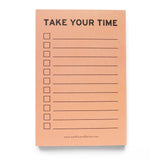 Vertical rectangle notepad with peach background, reading TAKE YOUR TIME in black text at the top, with left justified check boxes and lines to write on to the right. The brand's website is listed small at the bottom of the notepad: www.wordforwordfactory.com