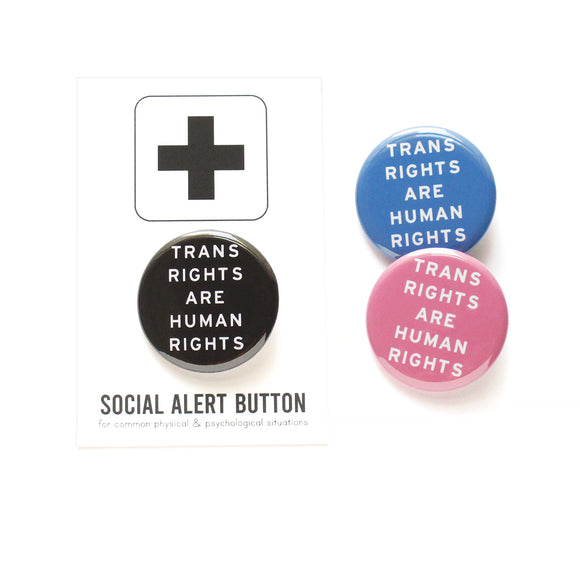 Three round pinback buttons that read TRANS RIGHTS ARE HUMAN RIGHTS. One is black, one is pink and one light blue, all have white text. The black button is on a Social Alert Button branded backing card.