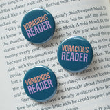 Three round teal colored pinback buttons which read Voracious Reader in sans serif font. Voracious is in peach, Reader is a larger word and in lavender. The button is on a type written page of a book.