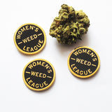 Three round WOMEN'S WEED LEAGUE enamel pins next to a nugget of cannabis